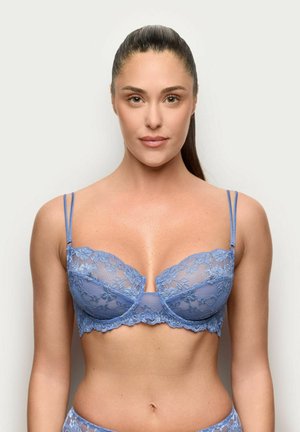 Donna con capelli lunghi e scuri raccolti in una coda di cavallo liscia, che indossa un completo intimo coordinato in pizzo azzurro chiaro, in piedi davanti a uno sfondo uniforme.