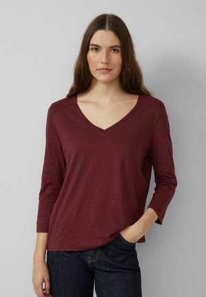Jeune femme aux longs cheveux bruns, portant un t-shirt à manches longues en vichy bordeaux et un jean foncé, avec une main dans la poche.