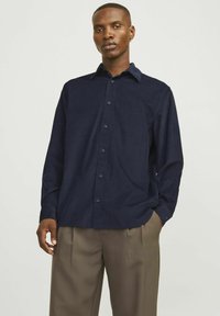 Camicia button-up blu navy realizzata in tessuto morbido, con colletto classico e maniche lunghe, abbinata a pantaloni chiari plissettati.