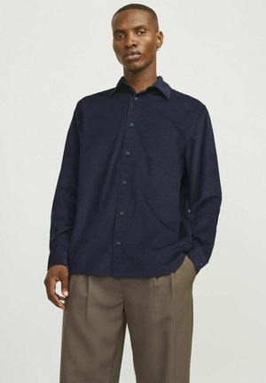 Camicia button-up blu navy realizzata in tessuto morbido, con colletto classico e maniche lunghe, abbinata a pantaloni chiari plissettati.