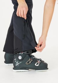 Zwarte skibroek met een rits en klittenbanddetails bij de enkel, gedragen over zwarte skiskchoenen met metallic gespen en textuurpatronen.
