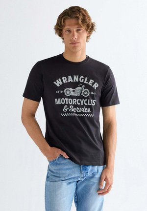 Młody mężczyzna w czarnej koszulce Wrangler z grafiką motocykla i jasnoniebieskich dżinsach, stojący na jednolitym białym tle.