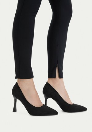 Nogi w czarnych dopasowanych spodniach z zamkami na kostkach, połączone z czarnymi szpilkami o wąskich obcasach i spiczastych czubkach na białym tle.