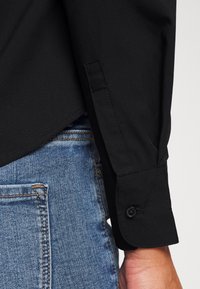 Camisa negra con puños abotonados y manga larga, metida dentro de unos vaqueros de denim azul, con una textura suave y un corte ajustado.