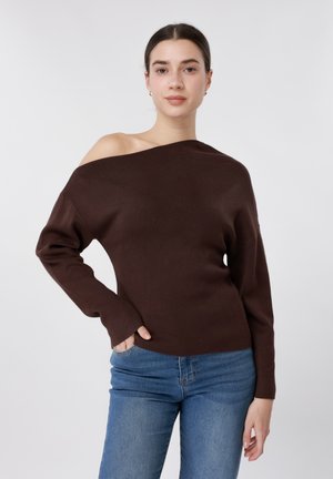 Γυναίκα που φοράει ένα σκούρο καφέ πουλόβερ έξω από τον ώμο και μπλε τζιν, όρθια με το ένα χέρι στην τσέπη, με φόντο ένα απλό φωτεινό υπόβαθρο.