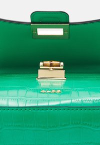 Sac à main en cuir vert à texture crocodile avec fermoir doré et détail en bambou, partiellement ouvert montrant le rabat intérieur et le logo de la marque.