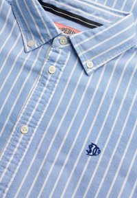 Camicia a righe verticali azzurra chiara e bianca con bottoni, colletto abbottonato e piccolo logo ricamato blu scuro sul petto.