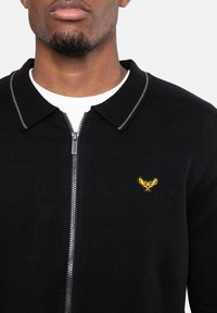Homme portant un pull zippé noir avec un petit emblème jaune brodé sur le côté gauche de la poitrine, par-dessus un t-shirt blanc à col rond.