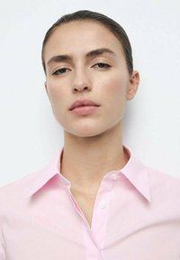 Cămașă roz cu nasturi, cu guler întins, realizată dintr-un material neted. Are un singur nasture la guler și elemente de design minimaliste.