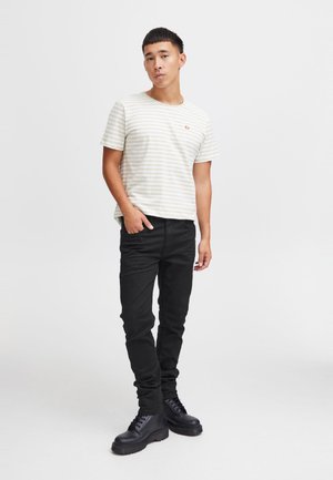 Beżowo-biała koszula w paski z krótkim rękawem, czarne dopasowane dżinsy i czarne buty. Na koszuli znajduje się małe logo na piersi.