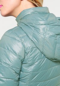 Kvinna i ljusblå vadderad jacka med huva, sedd från baksidan och sidan, med blont hår och klar rosa läppstift.