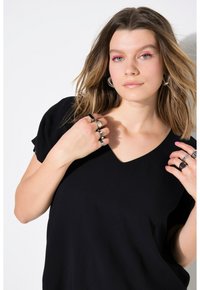 Jonge vrouw met schouderlang haar, gekleed in een zwarte V-hals top, meerdere zilveren ringen, ringoorbellen en roze eyeliner.
