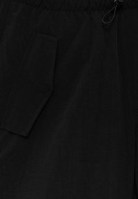 Gros plan sur un tissu noir montrant une poche texturée avec un rabat angulaire et un petit détail de boucle près du bord supérieur.