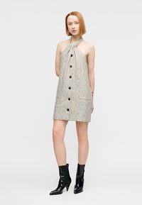 Robe sans manches beige texturée avec un col noué, des boutons noirs sur le devant et des poches latérales. Associée à des bottes à talons noires.