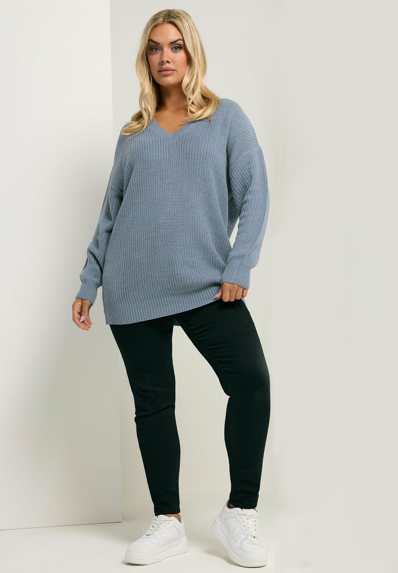 Jasnoniebieski sweter z dzianiny z dekoltem w serek i długimi rękawami, w połączeniu z czarnymi dopasowanymi spodniami i białymi trampkami. Miękka tekstura, luźny krój.