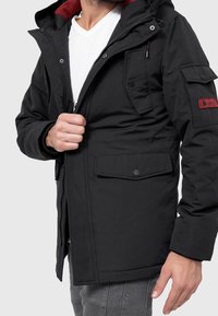 Svart jacka med frontdragkedja, röd innerfoder, flera fickor och en patch på ärmen. Tillverkad av vattenavvisande tyg.