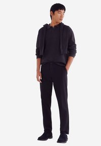 Schwarzer Pullover mit Kragen, kombiniert mit schwarzen Hosen und einer kurzen schwarzen Strickjacke. Das Outfit zeichnet sich durch eine matte Textur und ein minimalistisches Design aus.