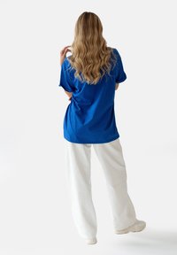 Niebieska, oversize koszulka z podwiniętymi rękawami, zestawiona z luźnymi białymi spodniami. Materiał wydaje się miękki, o gładkiej teksturze i prostym wzorze.