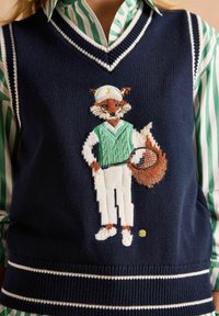 Tumes sinine kootud vest, millel on tikitud rebane tennisekohtumises, rohelise sviitri ja valgete pükste aktsentidega ning tennisereket ja pall.