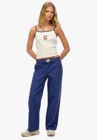 Hvid tanktop med sorte detaljer og "USA" tekst, parret med blå stribede bukser. Modellen bærer beige sko. Enkelt design og afslappet pasform.