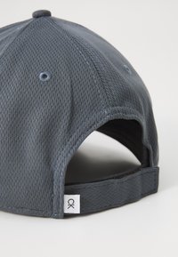 Grijze mesh pet met een ronde kroon en een verstelbare band. Bevat contrasterende stiksels en een klein logo label aan de zijkant.