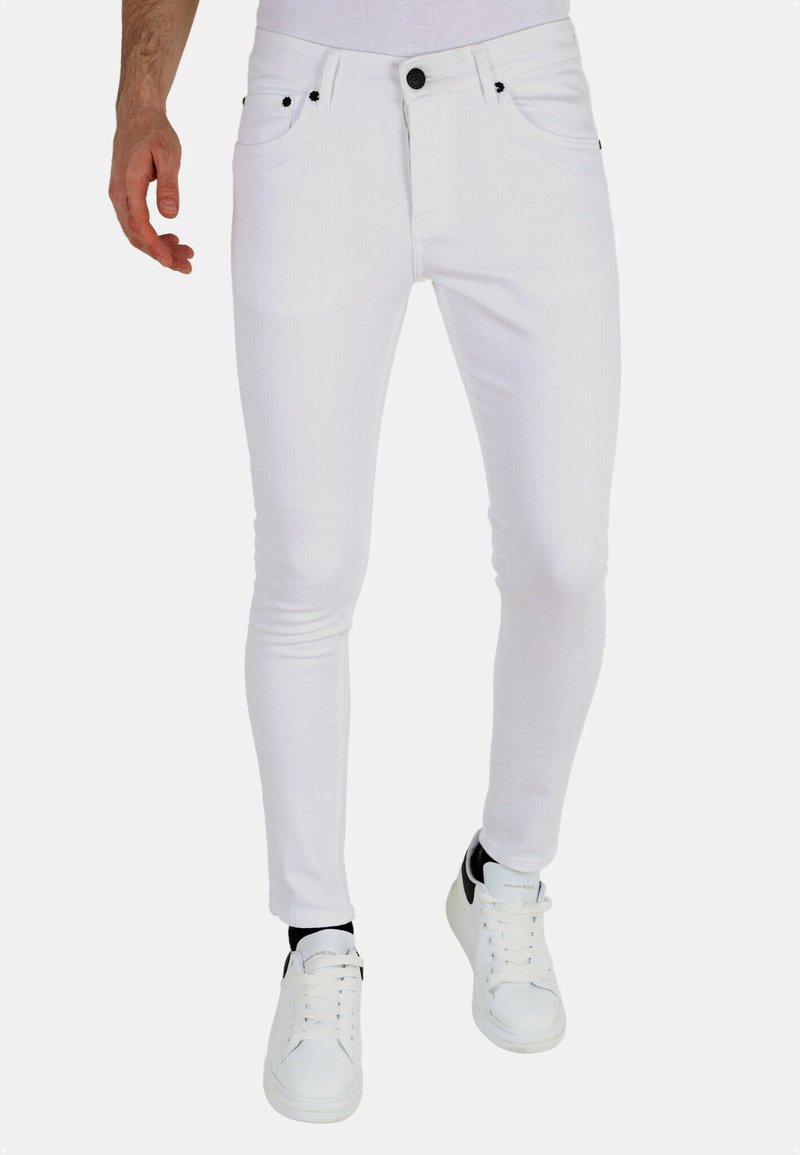 Witte slim-fit jeans met vijf zakken, contrasterende zwarte knopen en klinknagels, gecombineerd met witte sneakers met zwarte accenten.