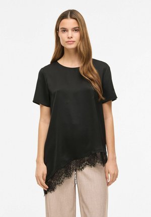 Femme aux longs cheveux bruns portant un haut noir à manches courtes avec un ourlet en dentelle asymétrique et un pantalon beige à jambes larges, debout devant un fond blanc.