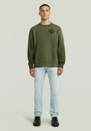 Jeune homme debout portant un sweat-shirt vert olive, un jean bleu clair et des chaussures noires sur un fond uni clair.