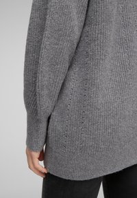 Gros plan sur la main d’une personne reposant sur le côté, vêtue d’un pull en maille grise texturée et d’un pantalon noir.