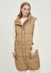 Gilet rembourré beige avec un col montant, des boutons-pression et deux poches avant. Présente une texture matelassée et un design sans manches.