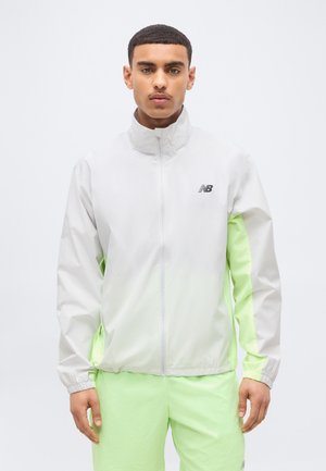 Jonge man die een wit en neon groen New Balance ritsjack draagt met bijpassende neon groene broek, staand tegen een effen lichte achtergrond.