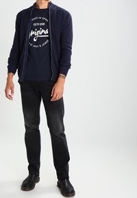 Marineblauer gerippter Cardigan über einem schwarzen Grafik-T-Shirt, kombiniert mit dunkelblauen Jeans und schwarzen Lederstiefeln. Das Outfit hat ein lässiges Design.