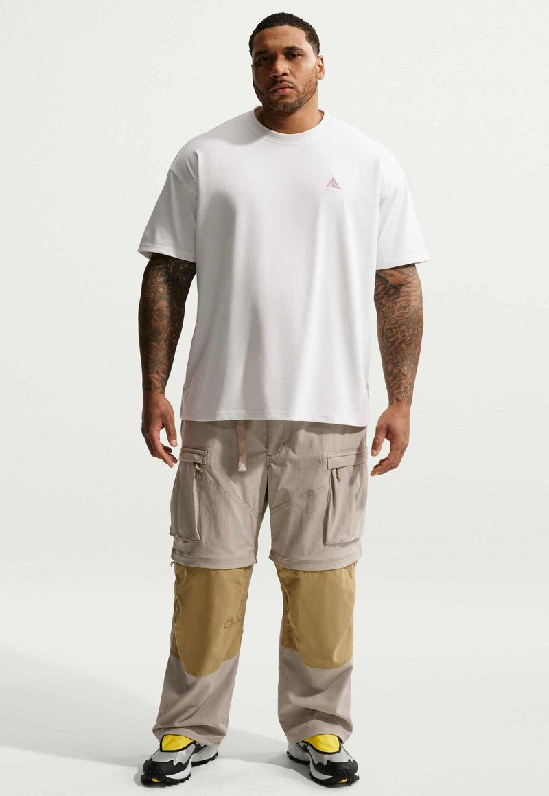 Homme aux bras tatoués portant une chemise blanche à manches courtes, un pantalon cargo beige avec des renforts aux genoux et des baskets avec des accents jaunes, debout sur un fond blanc.
