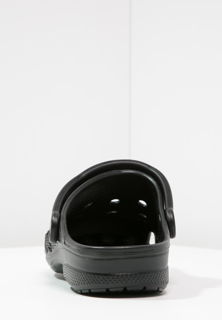 Zueco negro sin cierre con agujeros de ventilación y suela texturizada, visto desde atrás sobre una superficie blanca y fondo liso.