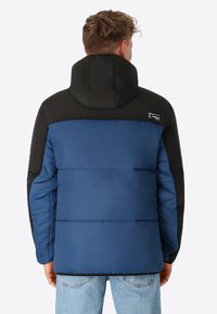 Gepolsterte Jacke mit blauem Körper, schwarzen Schultern und Kapuze. Sichtbare Nähte und Logodetail auf der linken Schulter. Weiche, glatte Textur.