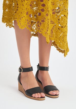 Czarne klapki na koturnie z gładką cholewką, odkrytym palcem, paskiem na kostkę z klamrą oraz plecioną teksturą na koturnie dla dodatkowego akcentu.