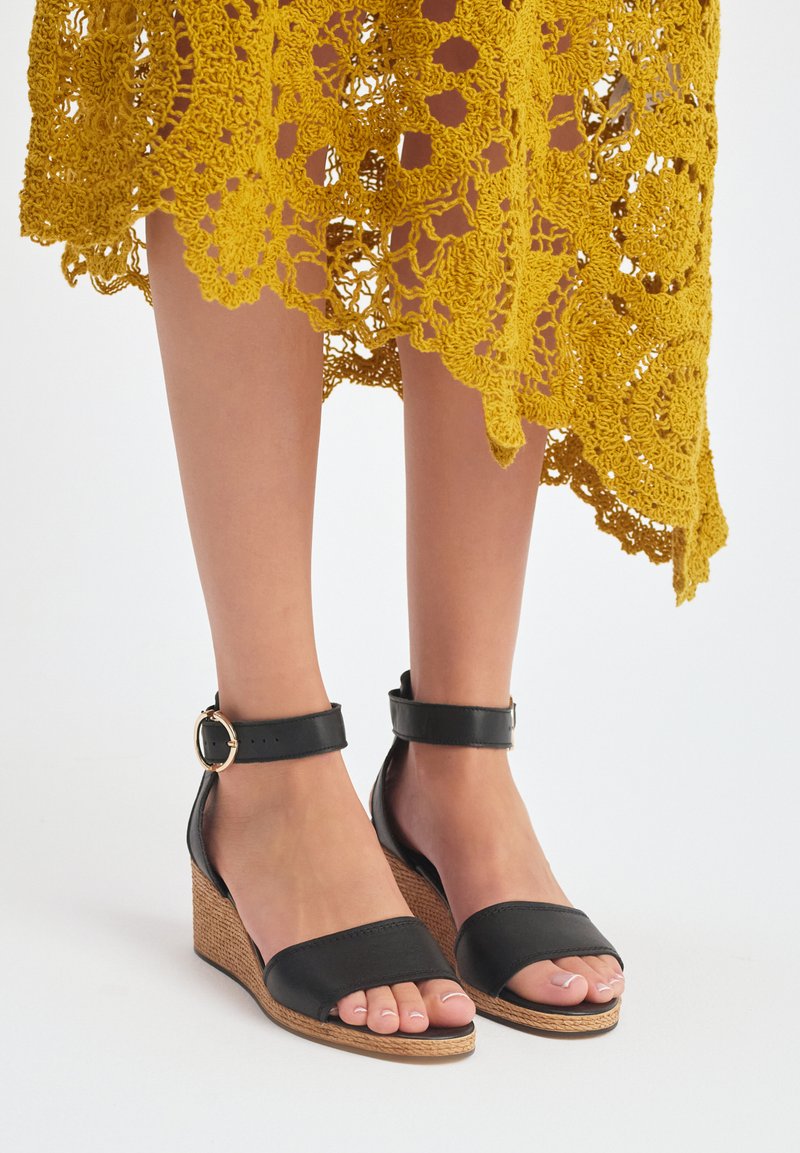 Czarne klapki na koturnie z gładką cholewką, odkrytym palcem, paskiem na kostkę z klamrą oraz plecioną teksturą na koturnie dla dodatkowego akcentu.