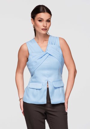 Femme aux cheveux foncés portant un haut sans manches bleu clair à empiècements plissés croisés et un pantalon noir, posant sur un fond uni.