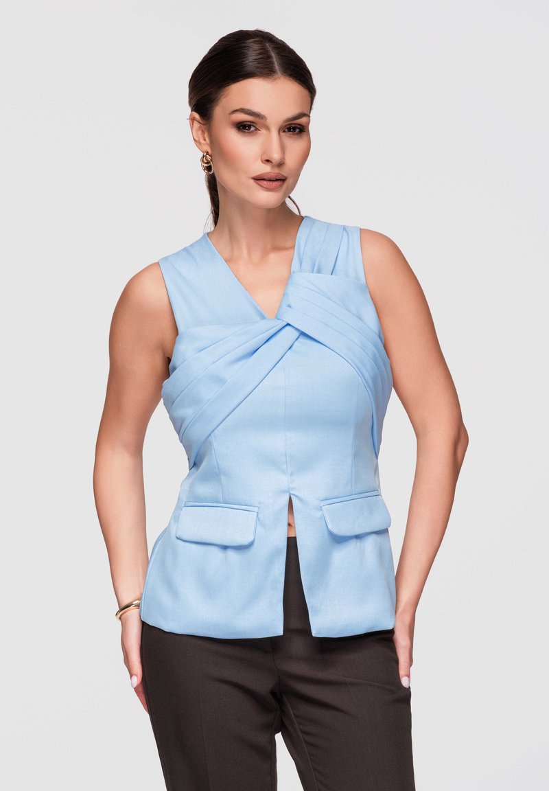 Femme aux cheveux foncés portant un haut sans manches bleu clair à empiècements plissés croisés et un pantalon noir, posant sur un fond uni.