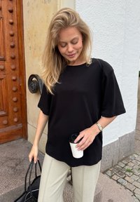 Juoda trumpomis rankovėmis marškinėliai, pagaminti iš švelnios medžiagos, derinami su šviesiai smėlinėmis kelnėmis. Ji laiko baltą puodelį su juodu dangteliu ir juodą maišelį.
