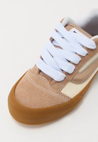 Beżowe zamszowe sneakersy z białymi sznurowadłami, gumową podeszwą i kremowym bocznym panelem. Zawierają detale szycia i zaokrąglony kształt palców.