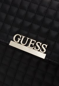 Mustakviltu faux nahasta valmistettu käsilaukku, jossa on hopeanvärinen "GUESS" logo ja tyylikkäitä metalliosia. Rakenteellinen pinta.