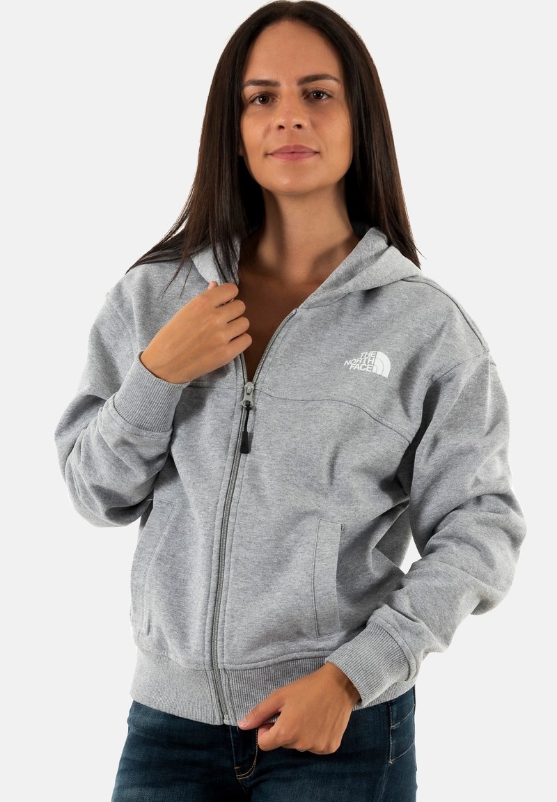 Sudadera gris con cremallera hecha de una tela suave. Presenta un bolsillo tipo canguro, puños acanalados y un logo en el pecho. La capucha está attached, lo que le da un aspecto informal.