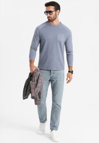 Vaaleansininen pitkähihais shirt, jossa on rintatasku, vaaleansinisiä farkkuja, valkoisia lenkkareita, kädessä ruudullinen flanellipaita ja aurinkolasit.