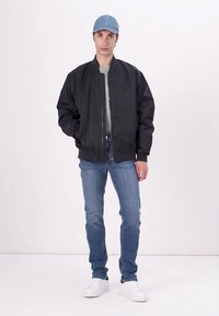 Veste bombardier noire avec fermeture éclair à l'avant, manches longues et poignets côtelés, associée à un jean bleu clair et des baskets blanches. Casquette bleue sur la tête.