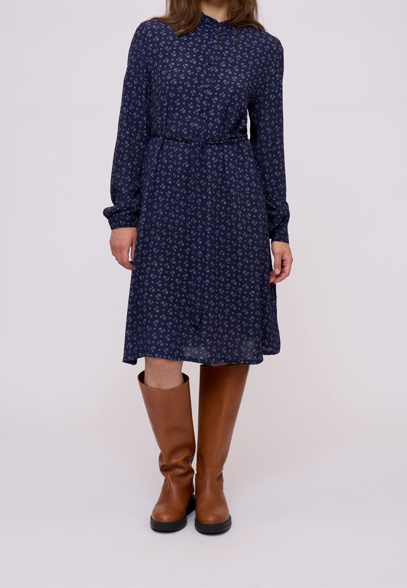 Femme portant une robe bleu marine à manches longues et motifs, nouée à la taille, avec des bottes marron jusqu'aux genoux, debout devant un fond clair et uni.