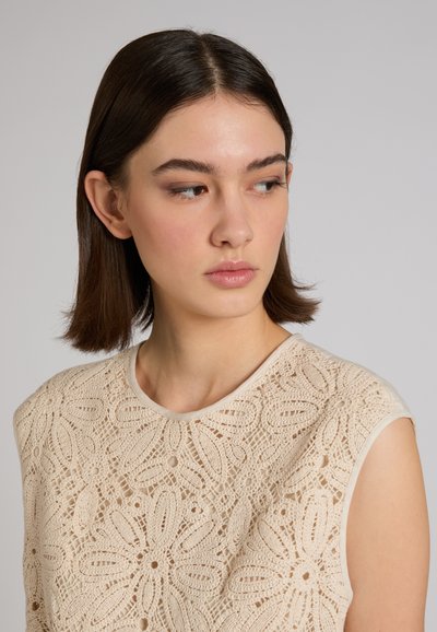 Młoda kobieta o prostych brązowych włosach, ubrana w beżowy, bezrękawnik z szydełkowanego materiału, patrząca w bok na jednolitym szarym tle.