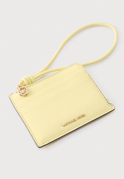 Porte-cartes en cuir texturé jaune pâle avec bracelet pour le poignet, logo Michael Kors doré et pendentif MK sur un fond uni clair.