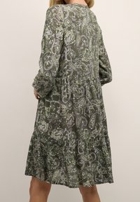 Groene jurk met paisleypatroon, lange mouwen, gelaagde ruches en een ronde halslijn. De stof heeft een zachte textuur en ingewikkelde details.