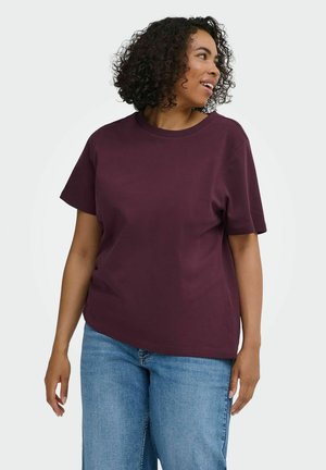 Femme aux cheveux bouclés portant un t-shirt violet à manches courtes et un jean bleu, regardant vers sa droite sur un fond uni.